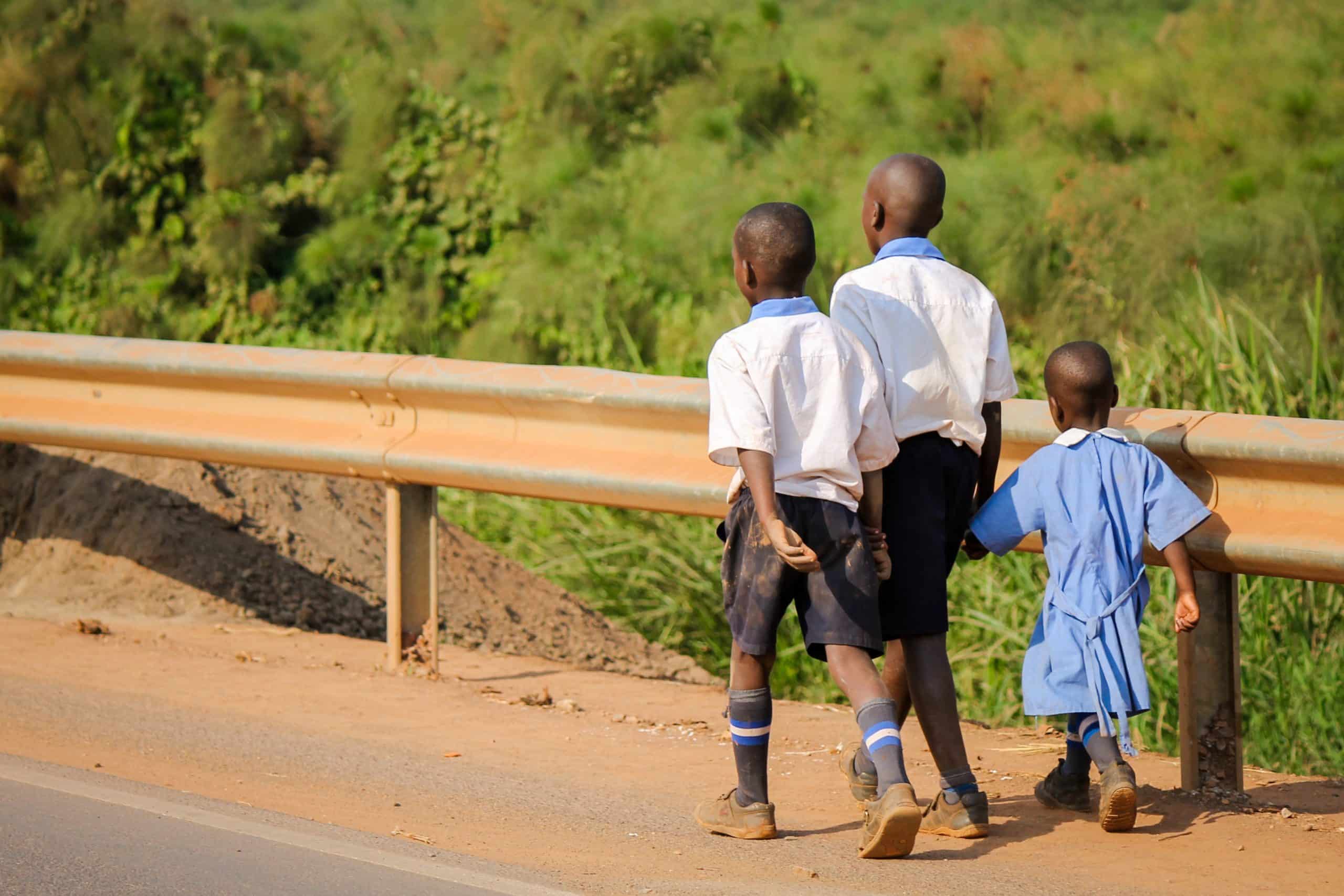 Three children in school uniforms walk along a road beside a guardrail, with greenery in the background.