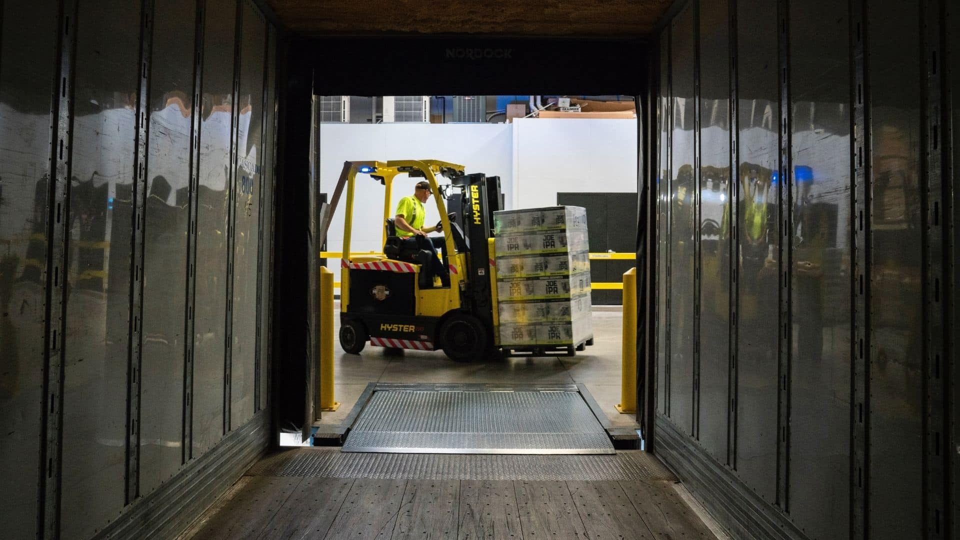A worker operates a forklift to move stacked pallets inside a warehouse, viewed from the interior of a loading dock or truck.